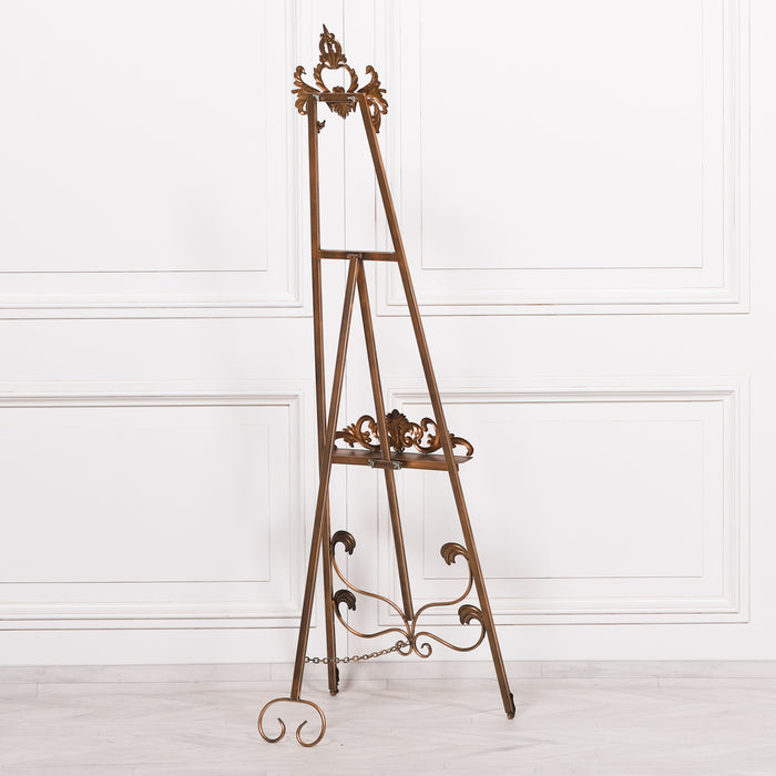 A 165cm tall French-style metal easel in antiqued gold finish with intricate details and an adjustable shelf.